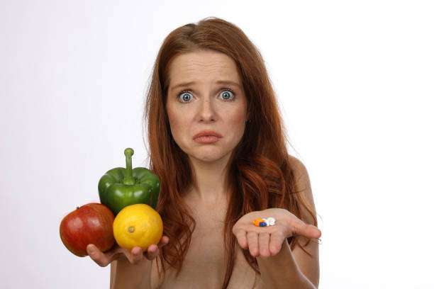 Young woman with red hair holds fruits, vegetables and tablets in front of her and is undecided