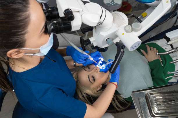 Two women dentist working with dental microscope. Doctor and his assistant in dental clinic. Dentist looking through microscope and holding dental probe and mirror. Modern dental clinic.