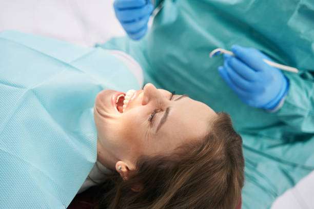 Female patient lying in dental chair and screaming dentist sitting beside woman and holding dental tools