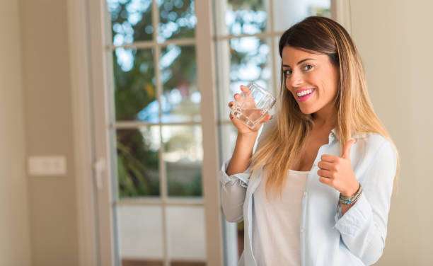 Beautiful young woman drinking a glass of water and doing ok sign with thumbs up at home. Lifestyle concept.