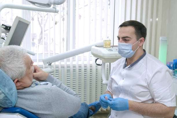 Elderly man sitting in dentist chair talks about his dental problems to doctor. Male dentist in dental room talking with senior patient and preparing for treatment. Healthcare, medicine and dentistry.