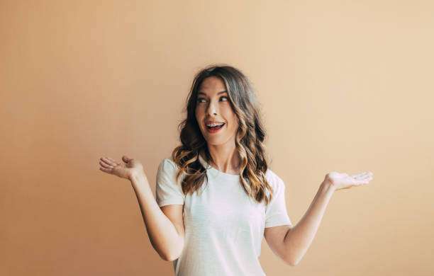 Surprised smiling young woman with braces on teeth demonstrating ads decisions choice with arms gesturing isolated on peach background.