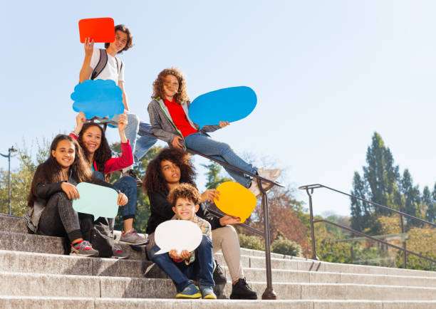 Group of teenage boys and girls sitting on the stairs outdoors, holding blanked speech bubbles