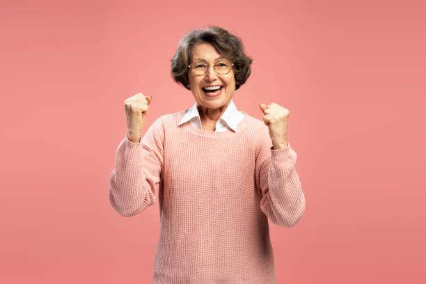 Portrait smiling funny senior woman, happy modern grandmother holding hands win something celebration success isolated on pink background. Good news, positive lifestyle concept